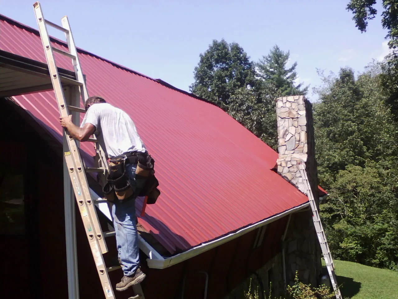 Roofer on a ladder beside a red metal roof and stone chimney — Soffit & Fascia Repair by Roofing Brothers in Wilmerding